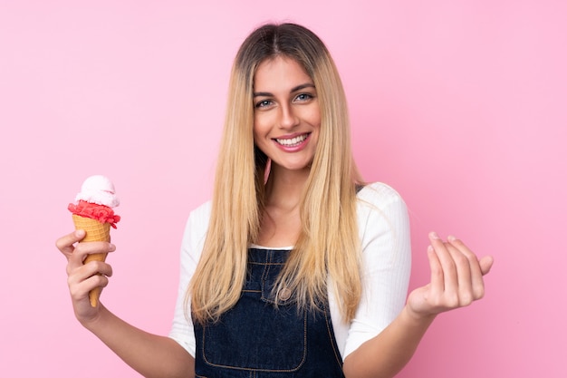 Young Uruguayan woman with a cornet ice cream over isolated wall pink wall inviting to come with hand. Happy that you came