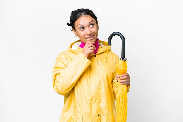 Young Uruguayan woman with rainproof coat and umbrella over isolated white background and looking up