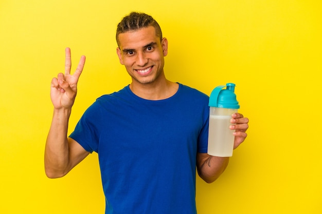 Young venezuelan man drinking a protein shake isolated on yellow background showing number two with fingers.