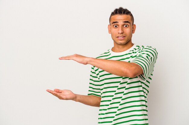 Young venezuelan man isolated on white background shocked and amazed holding a copy space between hands.