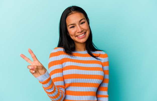 Young Venezuelan woman isolated on blue background showing victory sign and smiling broadly.