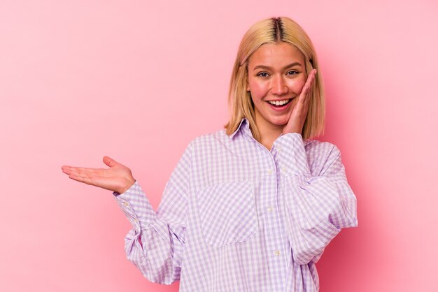 Young venezuelan woman isolated on pink background holds copy space on a palm, keep hand over cheek. Amazed and delighted.