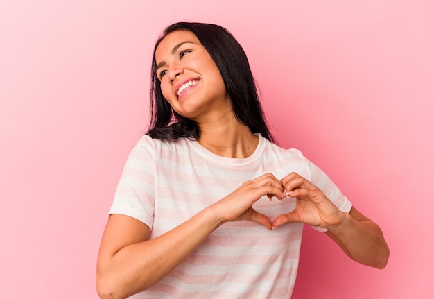 Young Venezuelan woman isolated on pink background smiling and showing a heart shape with hands.