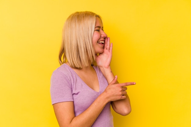 Young venezuelan woman isolated on yellow wall saying a gossip, pointing to side reporting something