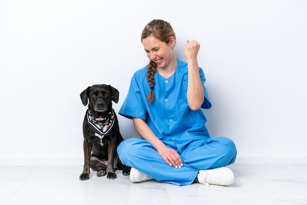 Young veterinarian woman with dog sitting on the floor isolated on white background celebrating a victory