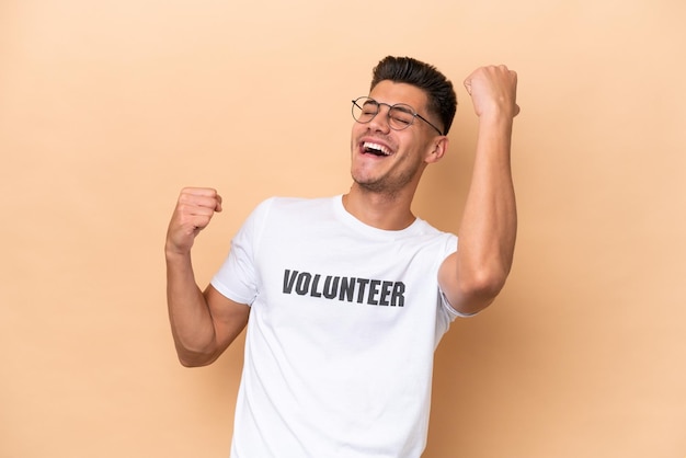 Young volunteer caucasian man isolated on beige background celebrating a victory