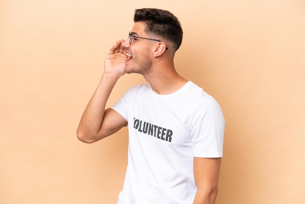 Young volunteer caucasian man isolated on beige background shouting with mouth wide open to the lateral