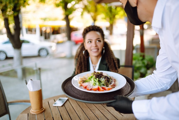 Young waiter in protective face mask and gloves with ordered meals ready to serving guest Covid2019