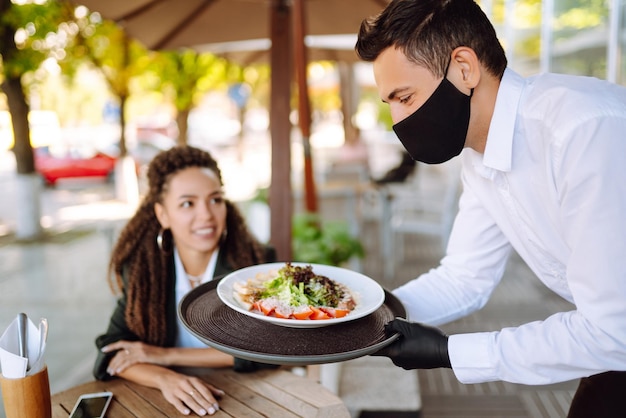 Young waiter in protective face mask and gloves with ordered meals ready to serving guest Covid2019