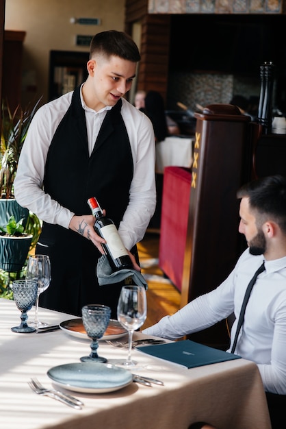 A young waiter in a stylish apron demonstrates and offers a fine wine to a client in a restaurant. Customer service.