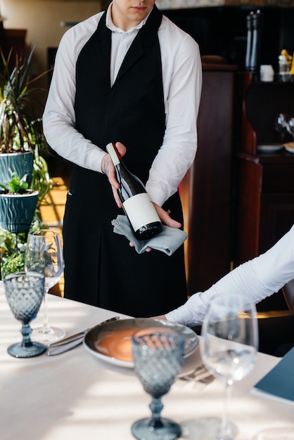 A young waiter in a stylish apron demonstrates and offers a fine wine to a client in a restaurant. Customer service.