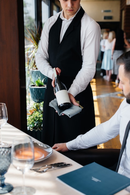 A young waiter in a stylish apron demonstrates and offers a fine wine to a client in a restaurant. Customer service.