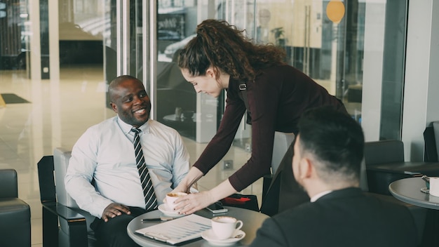 Young waitress bringing cup of coffee to businessmen in cafe