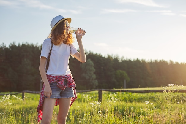 Young walking teen girl drinking water from a bottle. Background nature