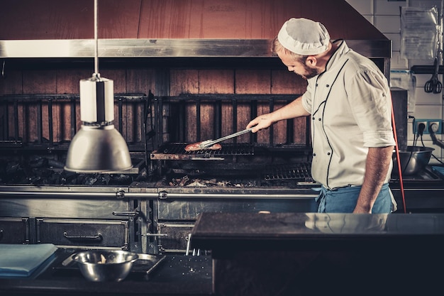 Young white chef in blue apron and hat standing near the brazier whith coals man cooking beef steak