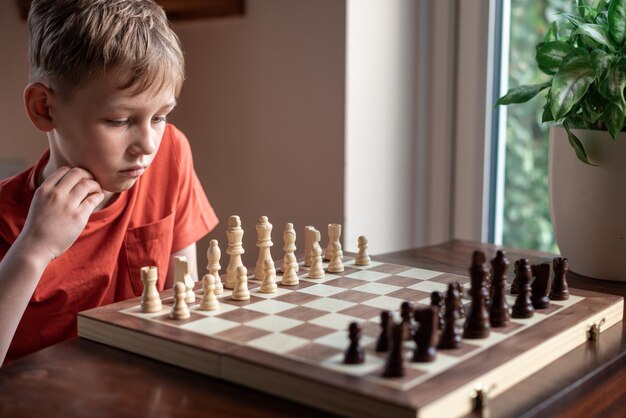 Young white child playing a game of chess on large chess board Chess board on table in front of school boy thinking of next move