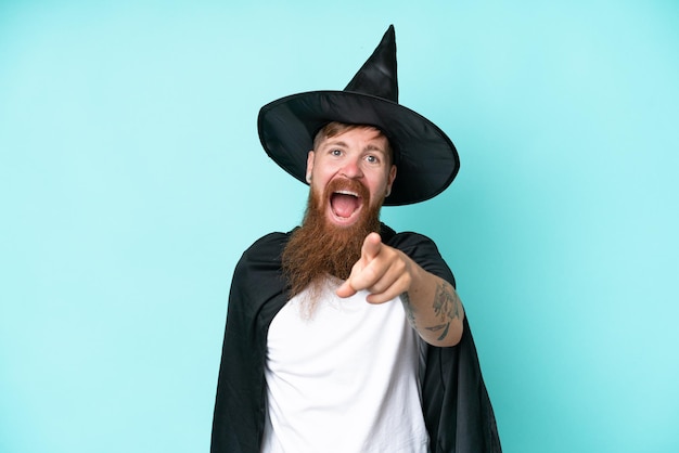 Young wizard in halloween isolated on blue background surprised and pointing front