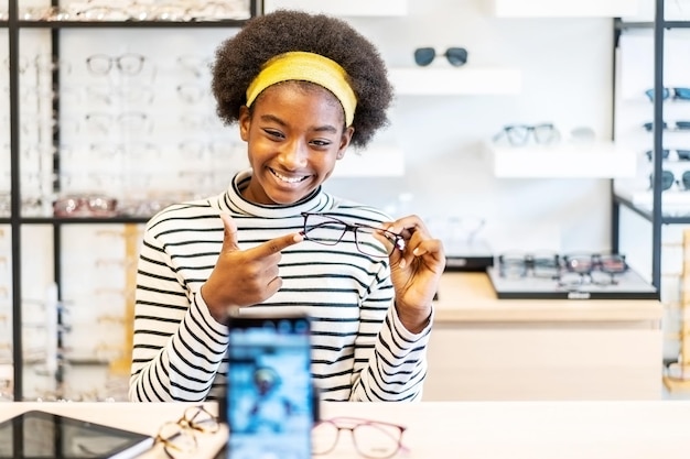 Young woman african american entrepreneur is fashion vlogger show glasses to cellphone and smiling selling it online live streaming in eyeglasses shop