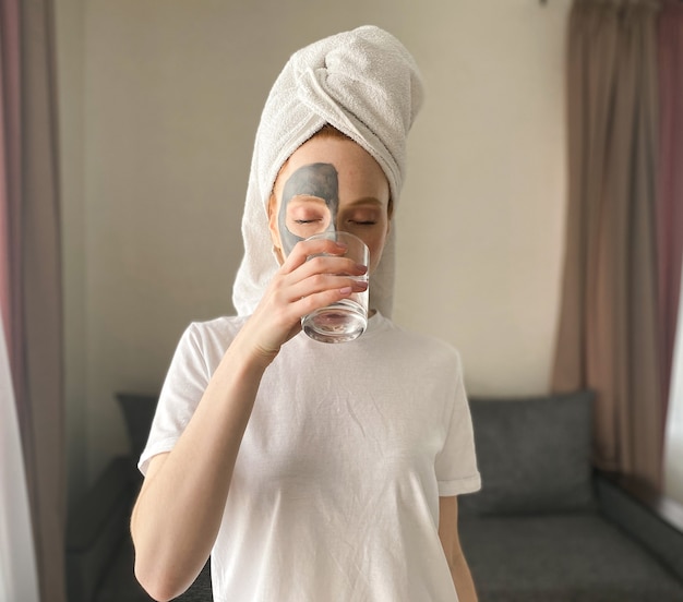 Young woman after a shower with a towel on her head, a clay mask on her face drinks water