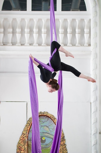 Young woman air gymnast in a black sportswear performs an exercise on aerial silk in a bright studio