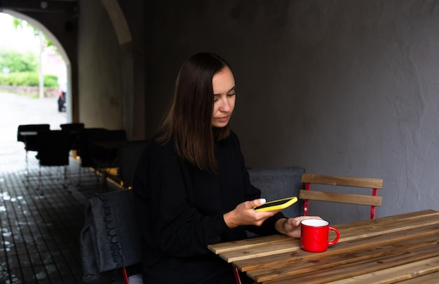 Young woman alone drinking in cafe on the terrace with a smartphone in her hands