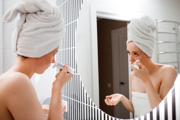 Young woman applying antiwrinkle cream standing behind mirror in home bathroom Cosmetology and beauty procedure Skin care after cleansing