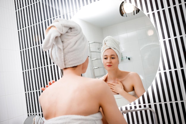 Young woman applying antiwrinkle cream standing behind mirror in home bathroom Cosmetology and beauty procedure Skin care after cleansing