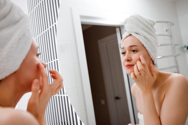 Young woman applying antiwrinkle cream standing behind mirror in home bathroom Cosmetology and beauty procedure Skin care after cleansing