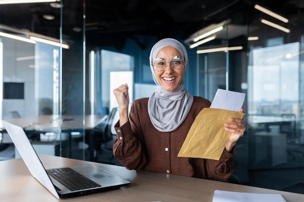 A young woman an arab student in a hijab sits at the table holds an envelope reads a letter received