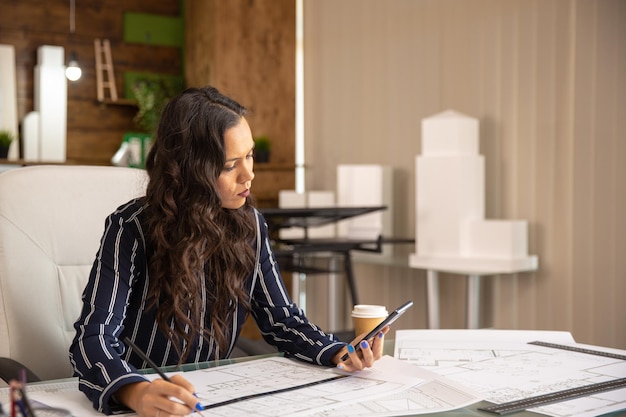 Young woman architect looking at phone and working on a project. blueprints new project