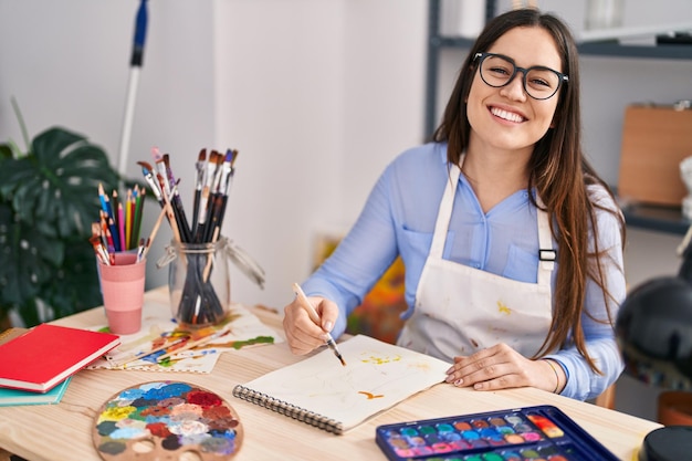 Young woman artist smiling confident drawing on notebook at art studio