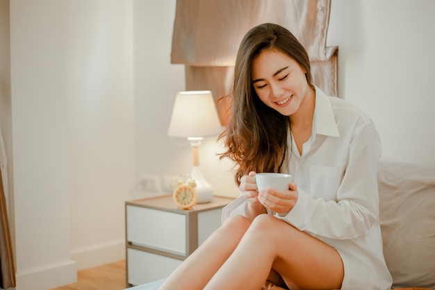 Young woman asia living at home relaxing and drinking cup of hot coffee in the bedroom on holiday. Asian, asia, relax, alone, technology, lifestyle concept.