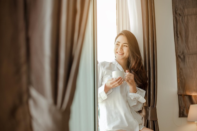 Young woman asia living at home relaxing and drinking cup of hot coffee in the bedroom on holiday. Asian, asia, relax, alone, technology, lifestyle concept.