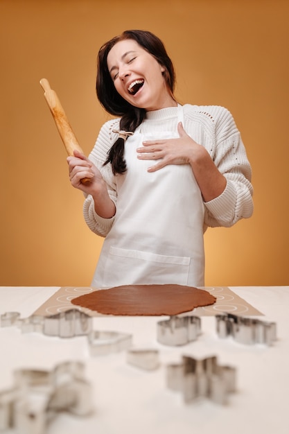 Young woman baker joyfully prepares dough for new years gingerbread on a yellow background in a whit...