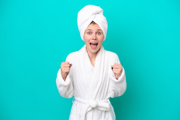 Young woman in a bathrobe isolated on blue background celebrating a victory in winner position