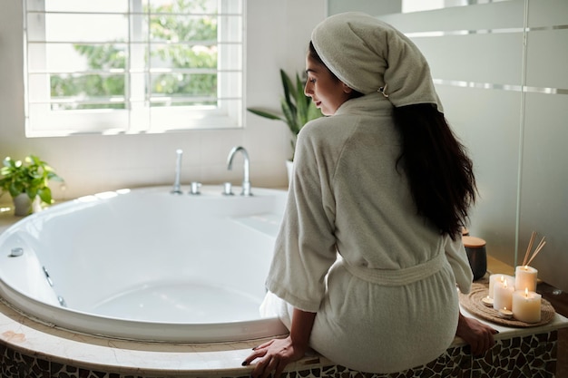 Young woman in bathrobe sitting next to bathtub spa day at home concept