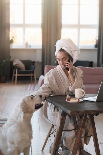 Young woman in bathrobe talking on mobile phone while sitting at the table with laptop at home