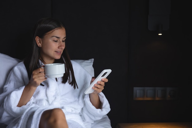 A young woman in a bathrobe with a cup of coffee and a smartphone in bed