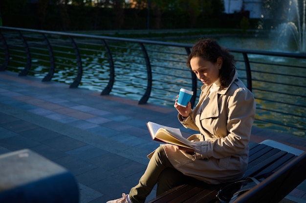 Young woman in beige trench coat sitting on a park bench on the lake background , drinking coffee or hot drink in recyclable takeaway paper cup and reading book, enjoying rest from digital gadgets