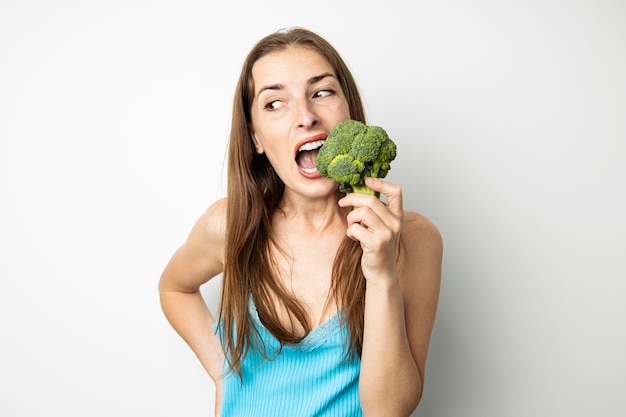 Young woman biting whole fresh green broccoli on white background