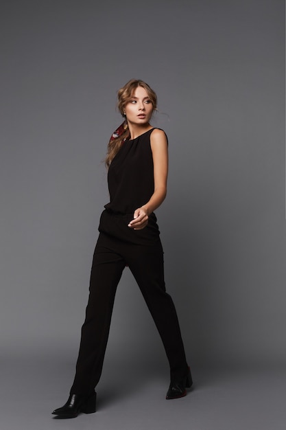 A young woman in a black casual outfit walking over the grey background. Model girl in a black suit on the grey background