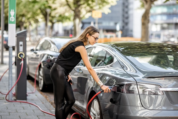 Young woman in black clothes putting connector into the electric car outdoors on the street in Rotterdam city