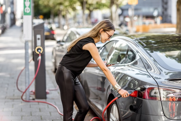 Young woman in black clothes putting connector into the electric car outdoors on the street in Rotterdam city