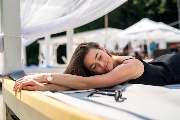 Young woman in black dress spending summer time and relaxing in fourposter bed near beach