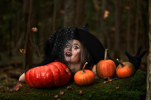 Young woman in black dress with witch hat and orange pumpkin placed around in the forest, Halloween concept. Horror theme.