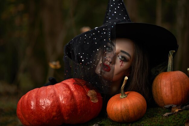 Young woman in black dress with witch hat and orange pumpkin placed around in the forest Halloween concept Horror theme