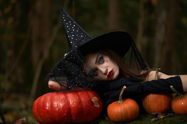 Young woman in black dress with witch hat and orange pumpkin placed around in the forest, Halloween concept. Horror theme.