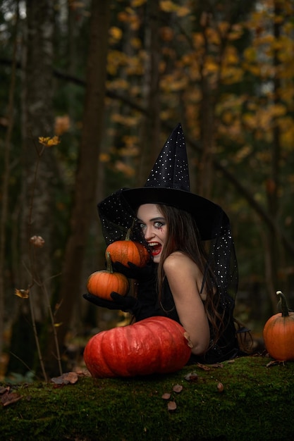 Young woman in black dress with witch hat and orange pumpkin placed around in the forest halloween