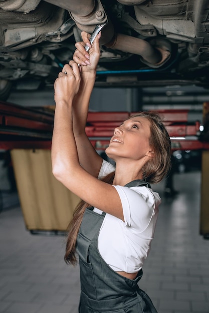 Young woman in black jumpsuit and white t-shirt unscrews the nut with a wrench under the car and smiles. female mechanic works under the underside of a car