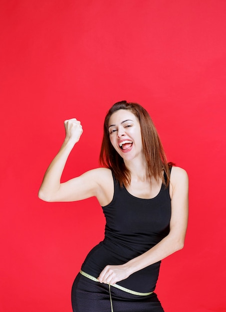 Young woman in black singlet holding a measuring tape, measuring her hips and feeling satisfied